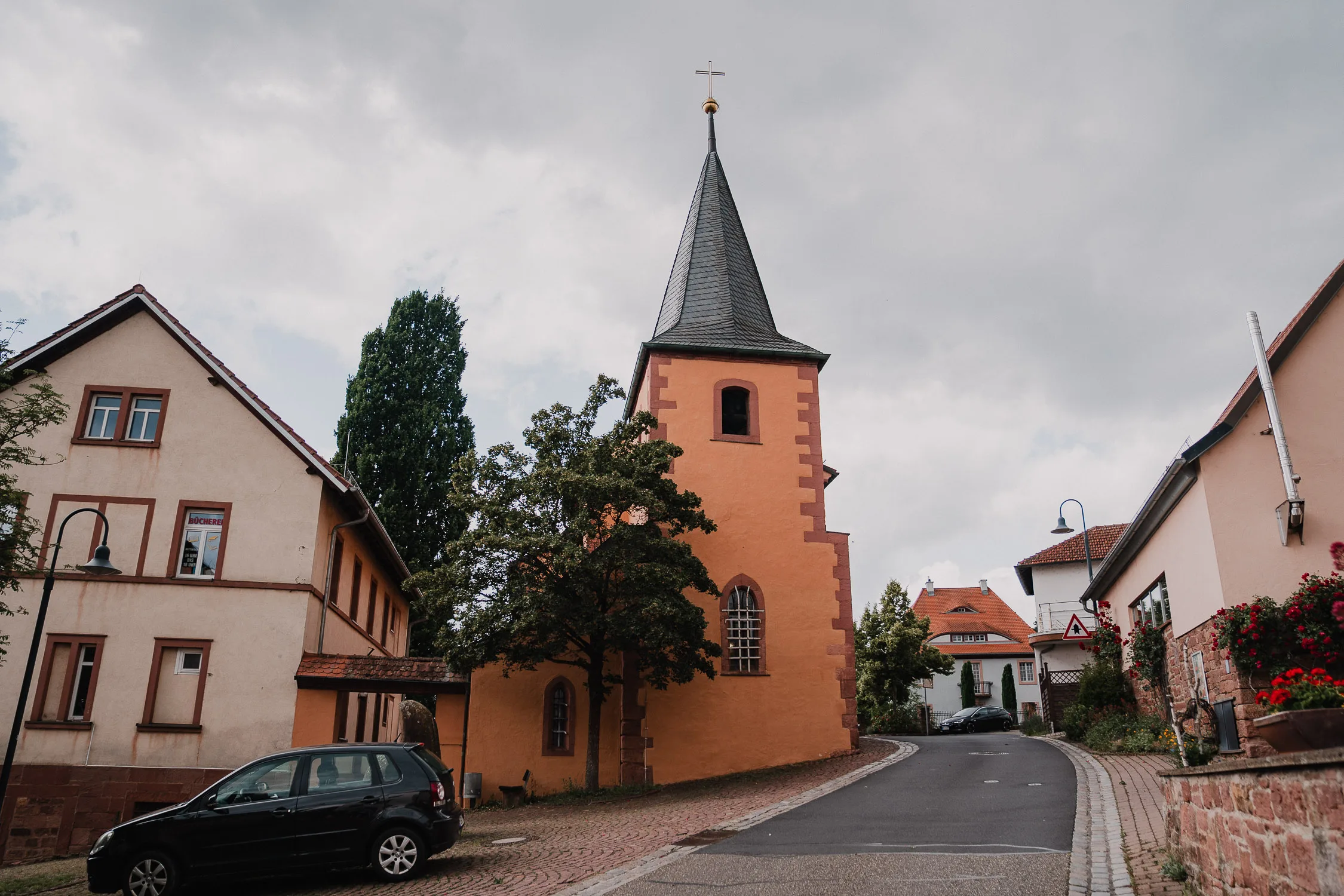 Eine schmale Straße schlängelt sich an einer kleinen orangefarbenen Kirche mit einem hohen Kirchturm vorbei, ein idealer Ort für Hochzeitsfotograf Würzburg, um Hochzeitsmomente einzufangen. Häuser und Grünflächen umgeben sie, während ein schwarzes Auto auf dem Kopfsteinpflaster ruht.