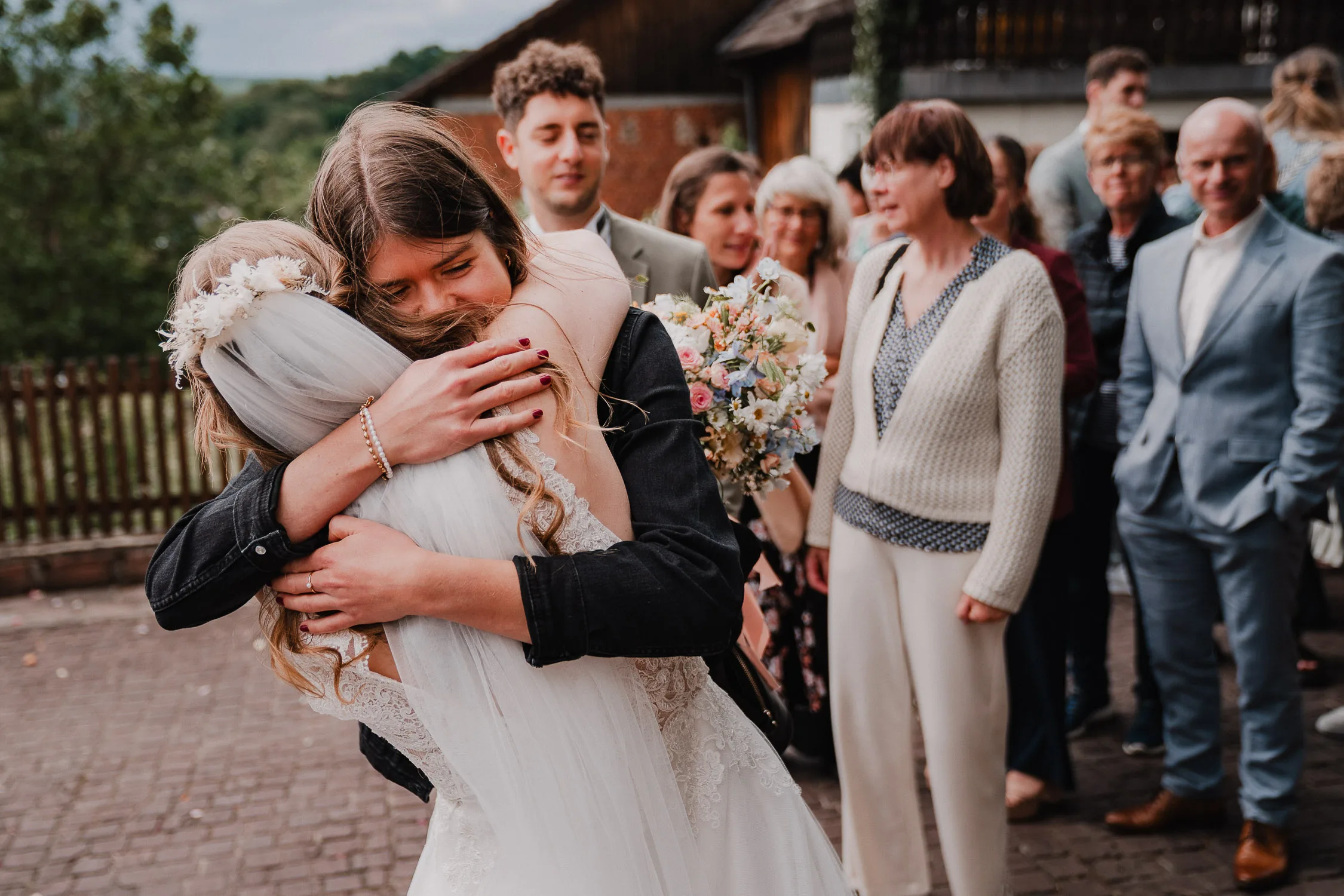 Eine Braut in einem weißen Kleid umarmt einen Gast mit langen Haaren, während andere lächelnde Gäste in der Nähe stehen, einige mit Blumen in der Hand, und die Freude der Hochzeit festhalten - perfekte Momente für jeden Hochzeitsfotografen Würzburg, um sie in Erinnerung zu behalten.