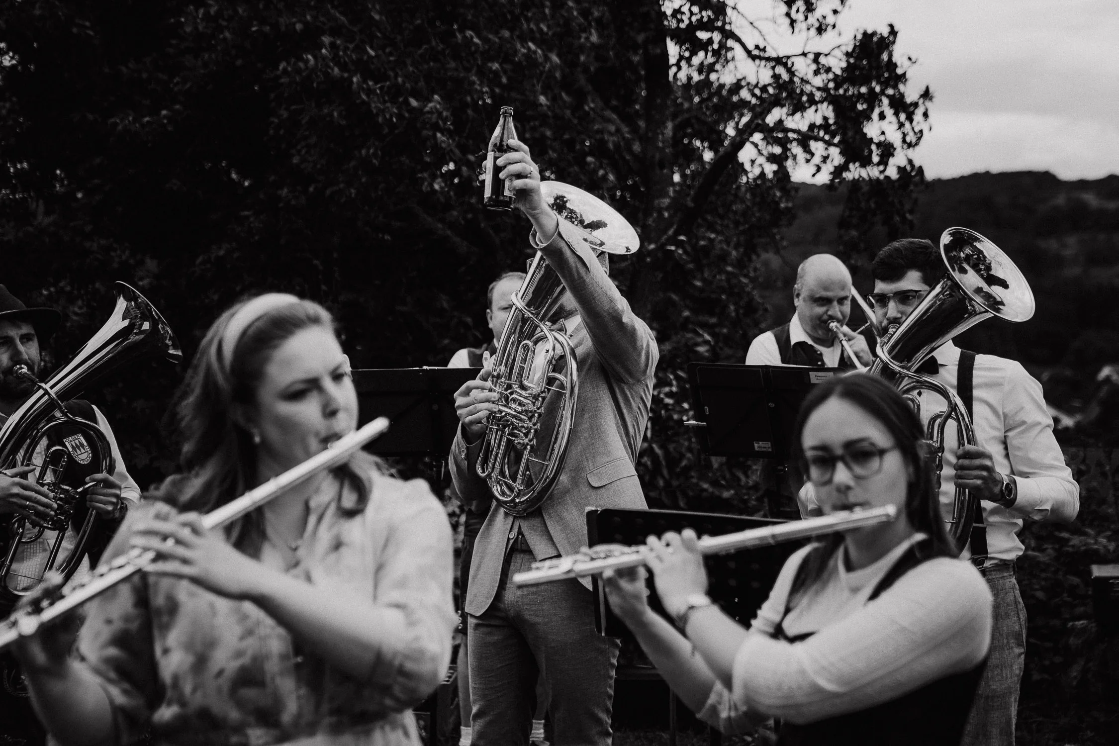 Ein Schwarz-Weiß-Foto einer Musikkapelle, die bei einer Hochzeit im Freien spielt. Zwei Personen im Vordergrund spielen Flöte, während hinter ihnen Blasmusiker stehen. Ein Musiker hebt zur Feier des Tages eine Flasche in die Höhe. Im Hintergrund sind Bäume und Hügel zu sehen.