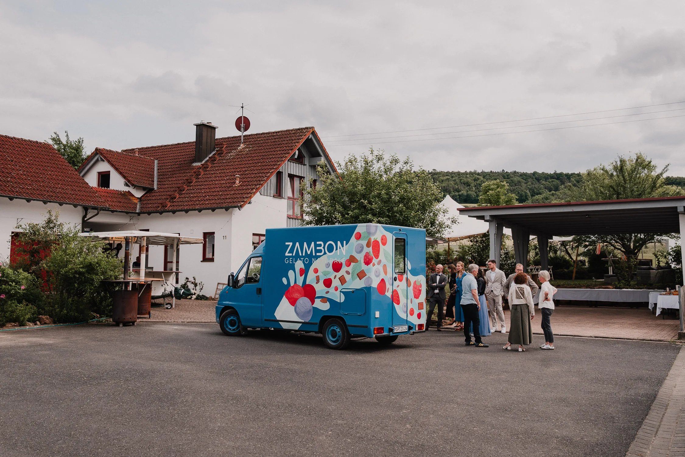 Eine Gruppe versammelt sich in der Nähe eines blauen Zamboni-Gelato-Trucks, möglicherweise auf einer Hochzeit, geparkt vor weißen Häusern mit roten Dächern und üppigem Grün. Der bedeckte Himmel gibt die Stimmung vor - perfekt für einen Hochzeitsfotografen Würzburg, um unvergessliche Momente festzuhalten. 