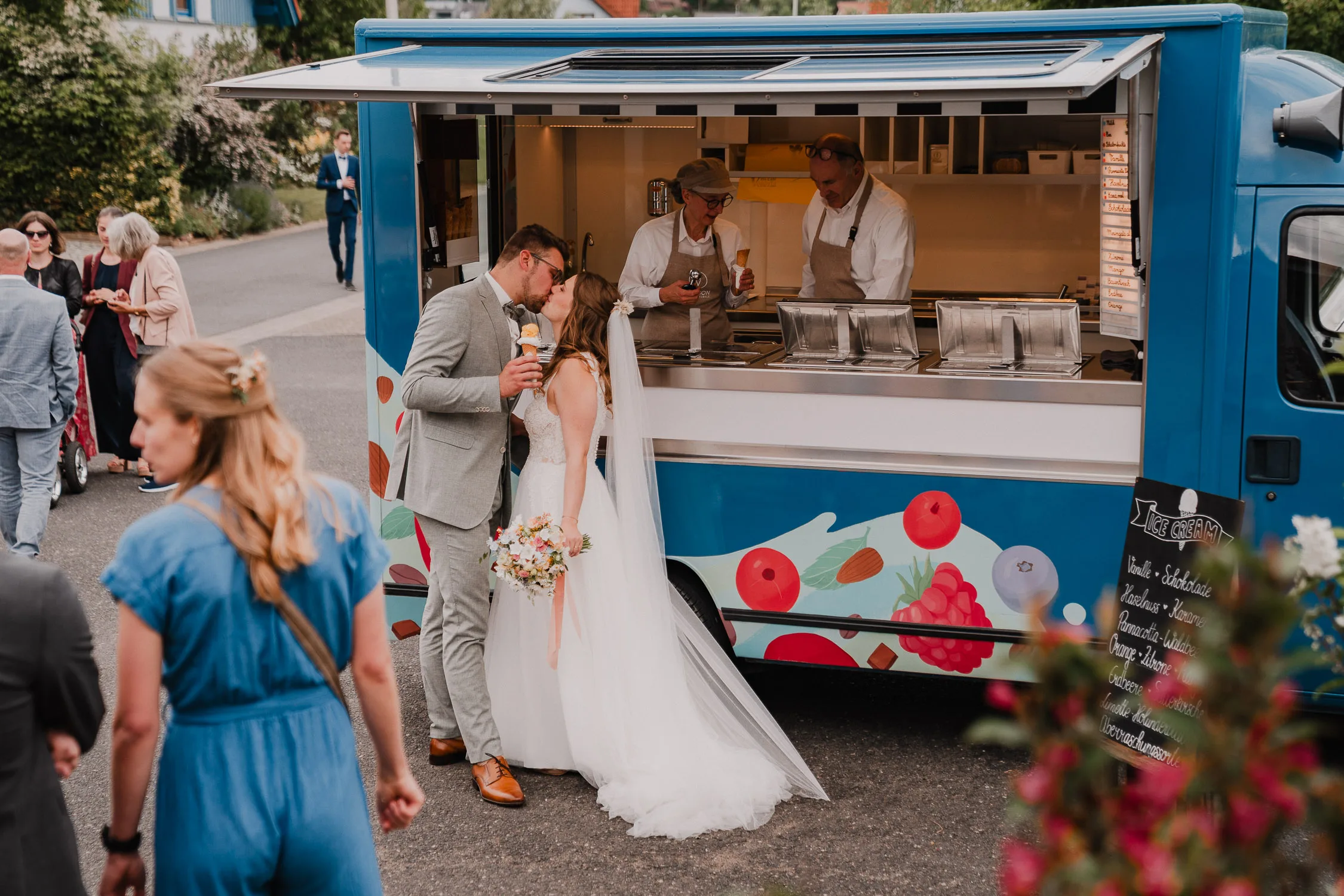 Braut und Bräutigam in Hochzeitskleidung küssen sich vor einem farbenfrohen Foodtruck auf ihrer Gartenhochzeit, während das Personal im Inneren das Essen serviert. Die Gäste mischen sich in der Nähe an einem sonnigen Tag im Freien, aufgenommen von Hochzeitsfotograf Würzburg.