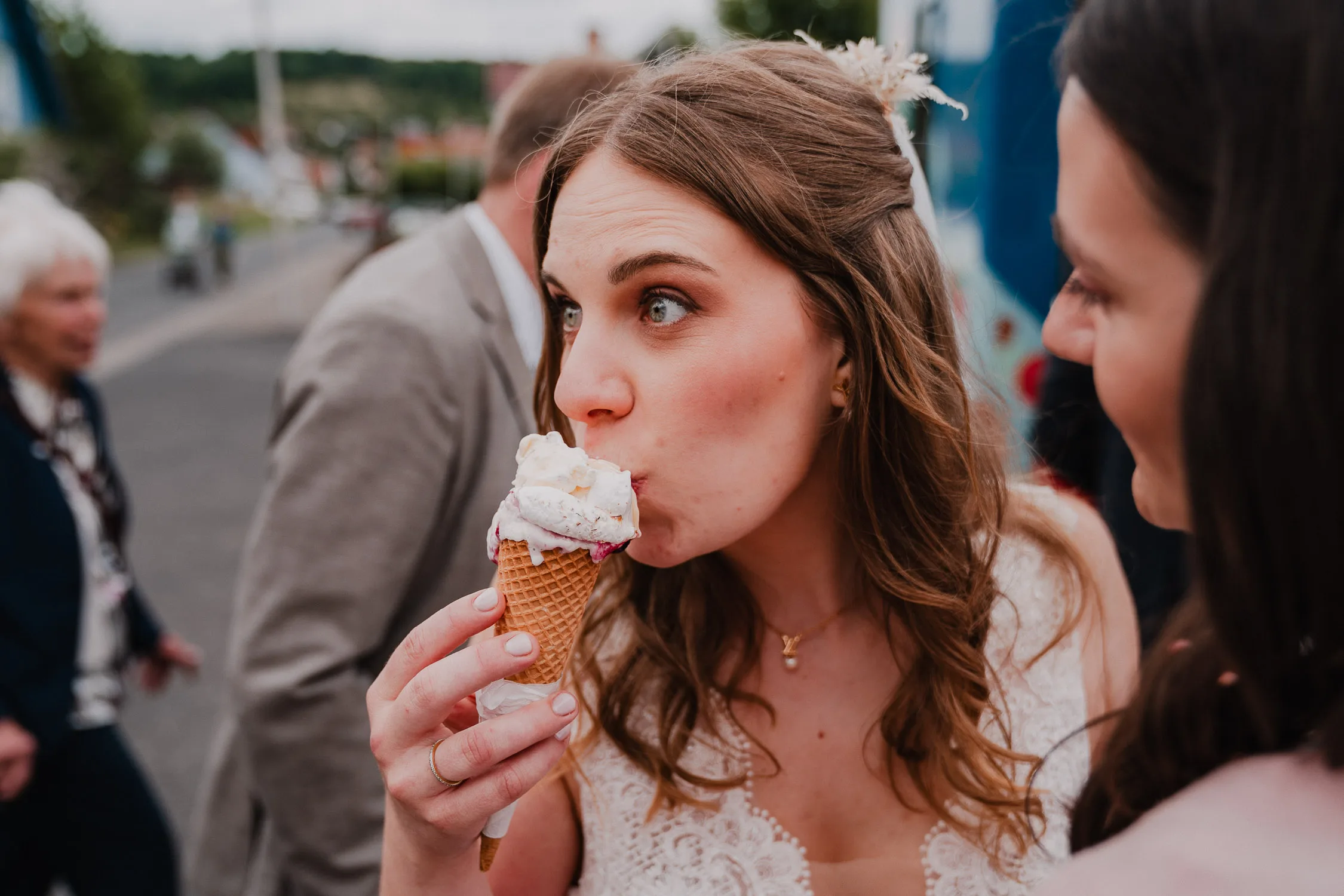 Eine Braut in einem weißen Kleid isst eine Eistüte im Freien und freut sich auf eine Gartenhochzeit. Andere Personen und der Fotograf sind im Hintergrund unscharf.