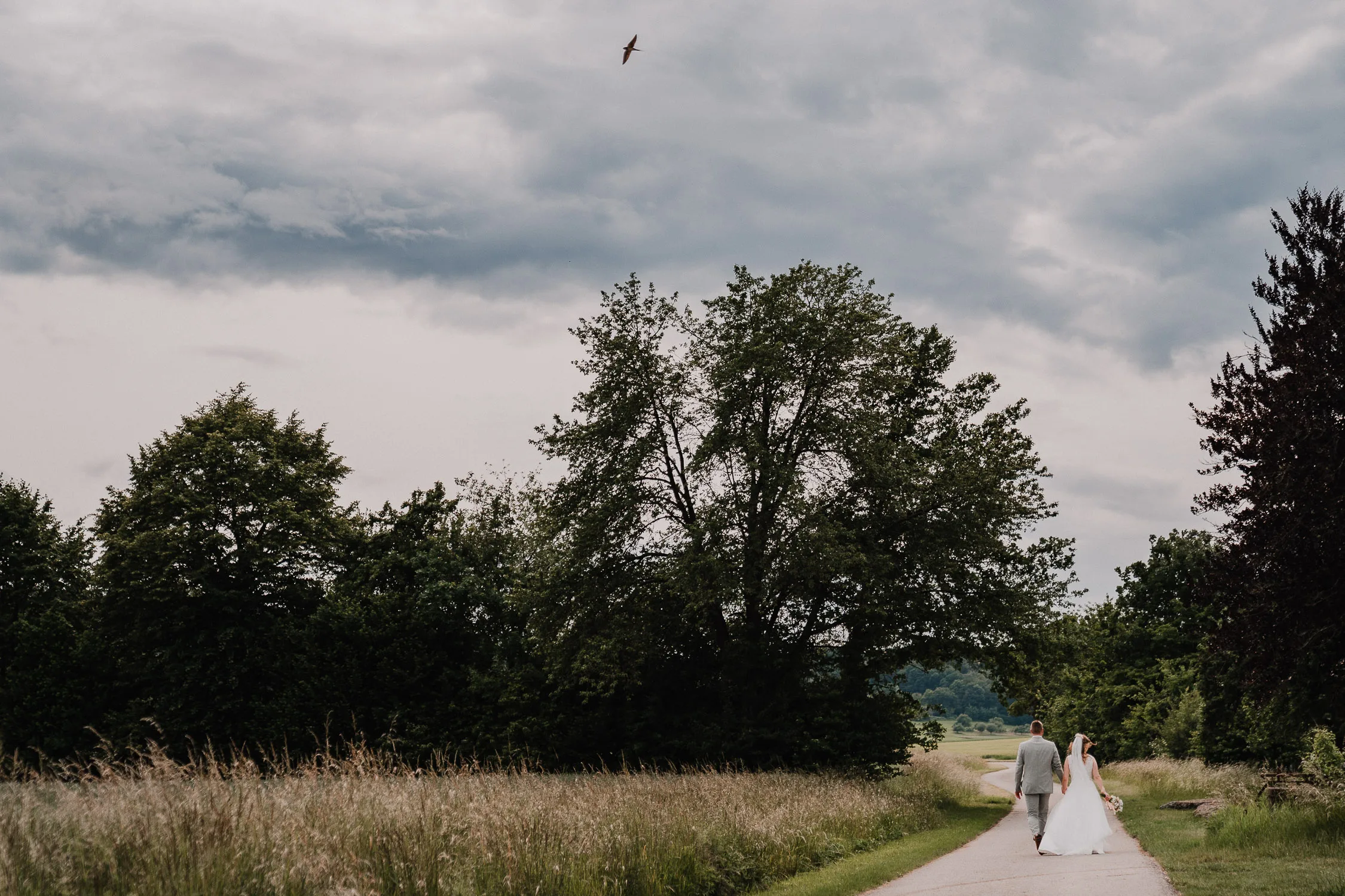 Braut und Bräutigam gehen Hand in Hand einen Feldweg entlang, umgeben von hohen Gräsern und Bäumen, unter einem wolkenverhangenen Himmel, über dem ein Vogel fliegt - der Zauber einer Gartenhochzeit aus der Sicht eines Hochzeitsfotografen Würzburg.