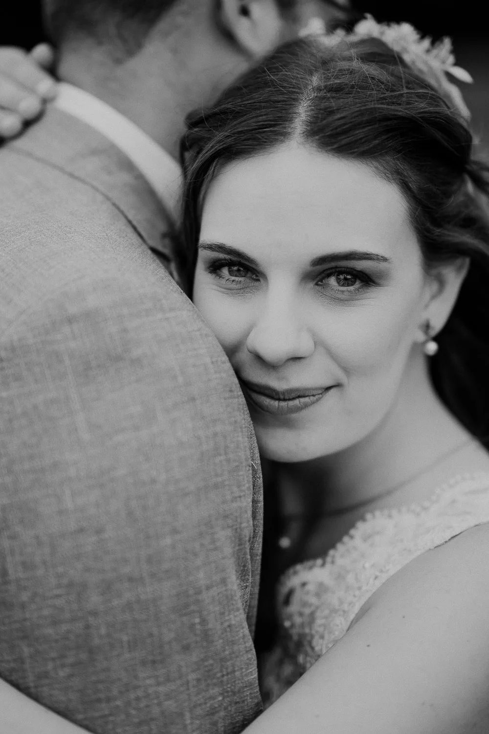 Eine Frau in einem Hochzeitskleid umarmt einen Mann im Anzug und blickt sanft in die Kamera. Das Schwarz-Weiß-Bild wurde von einem Hochzeitsfotografen aus Würzburg aufgenommen und hebt ihren heiteren Ausdruck und die zarten Details ihres Kleides hervor.