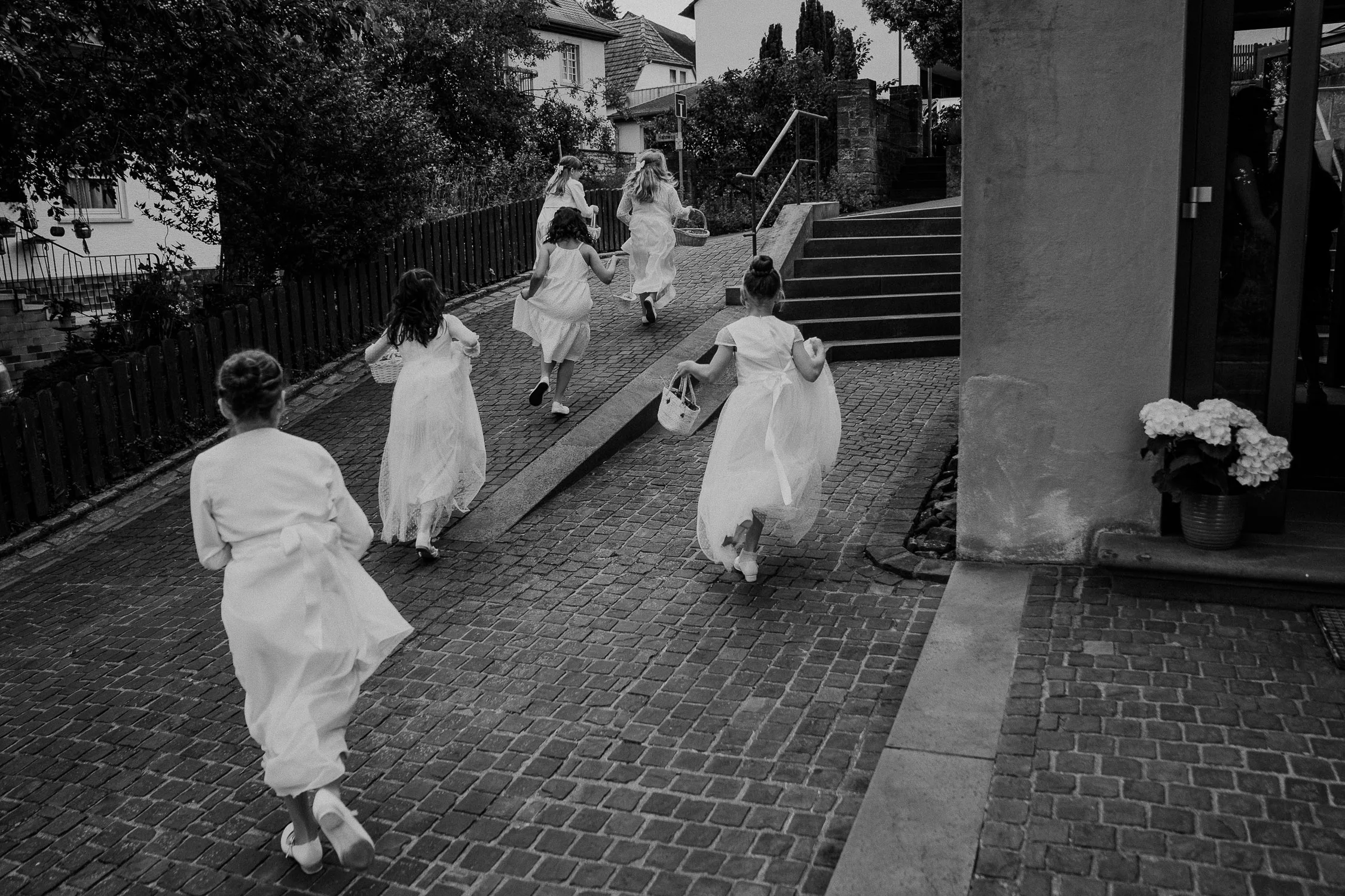 Brautmädchen in weißen Kleidern und Blüten zum Streuen rennen um die Kirche, als sie erfahren, dass die Braut angekommen ist. Eingafangen vom Hochzeitsfotografen aus Würzburg.