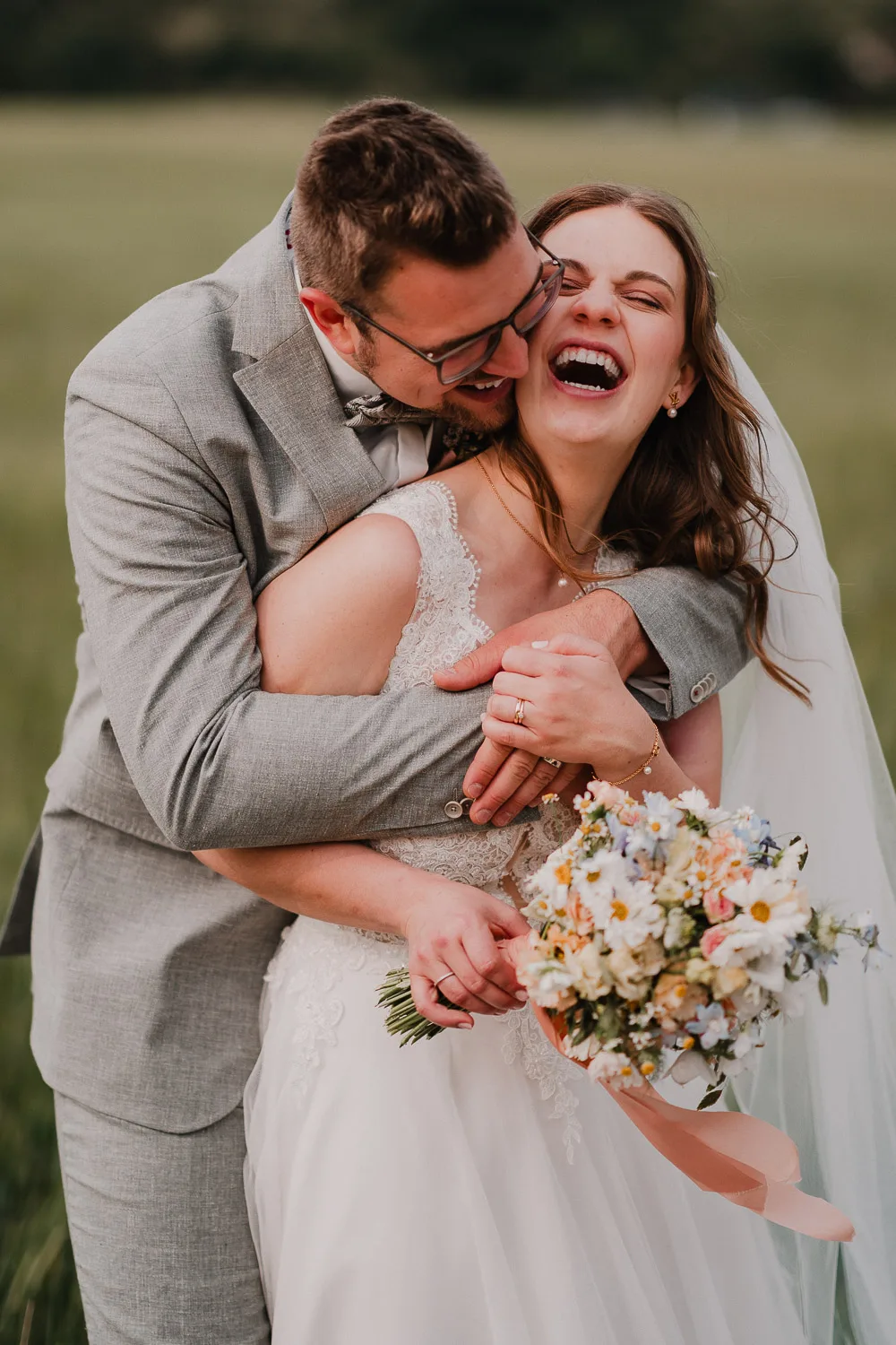 Ein Bräutigam in einem grauen Anzug umarmt seine lachende Braut in einem weißen Spitzenkleid, während sie ihren Blumenstrauß hält. Aufgenommen von einem Hochzeitsfotograf Würzburg, teilen sie einen fröhlichen Moment im Freien während ihrer Gartenhochzeit.