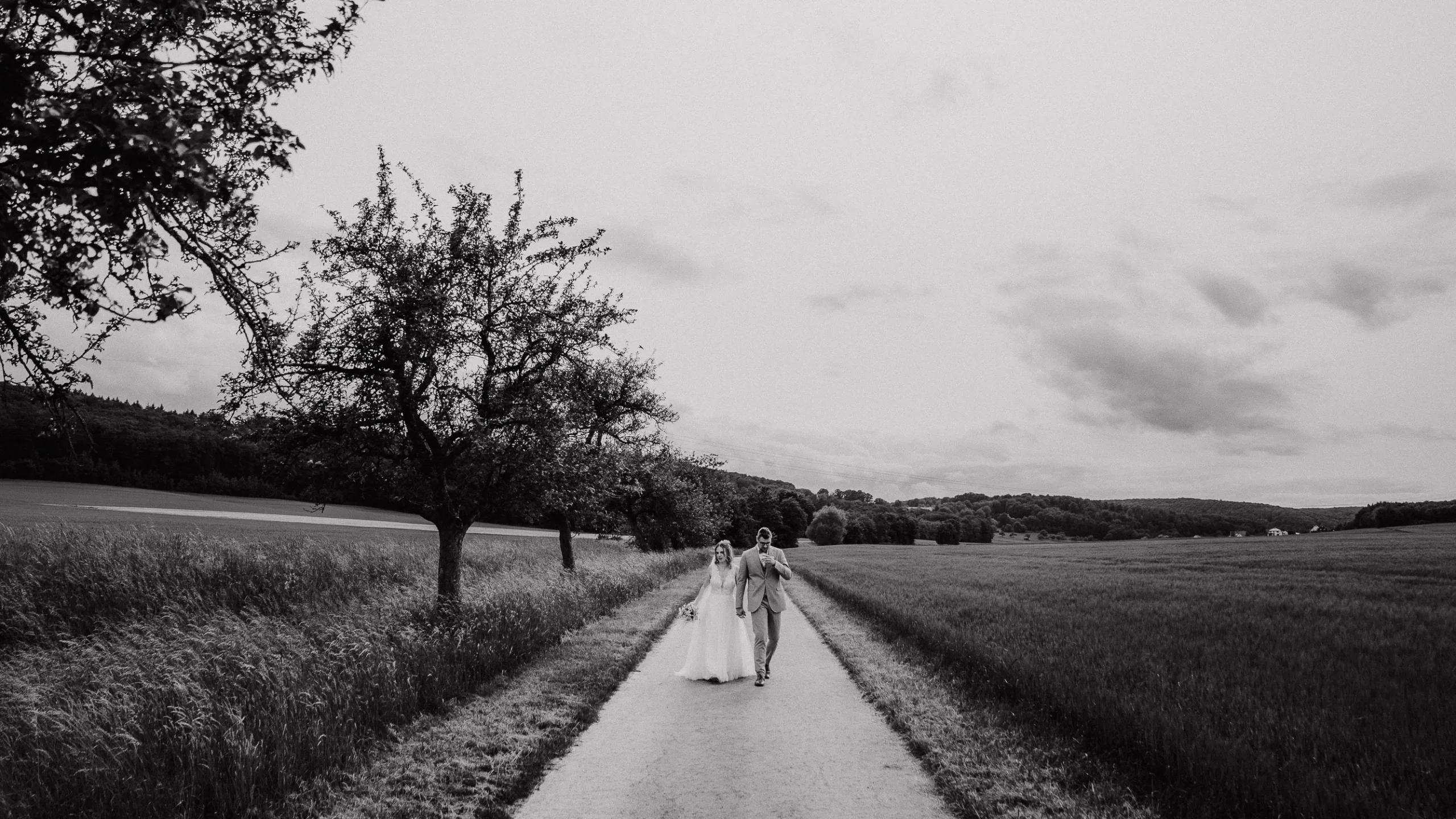 Eine Braut und ein Bräutigam gehen Hand in Hand einen von Bäumen und Feldern gesäumten Feldweg unter einem bewölkten Himmel entlang. Das Schwarz-Weiß-Foto, aufgenommen von einem Hochzeitsfotograf Würzburg, schafft eine zeitlose, romantische Atmosphäre.