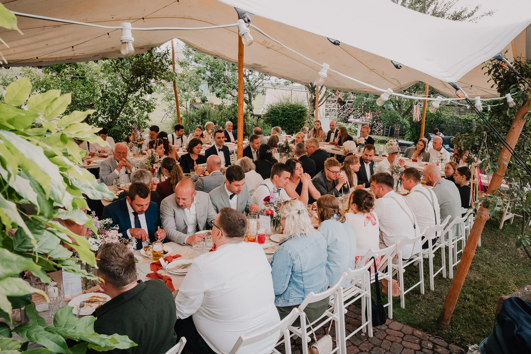 Eine große Gruppe von Menschen sitzt an langen Tischen unter einem weißen Zelt und genießt eine gemeinsame Mahlzeit bei einer Hochzeit im Freien, umgeben von Grün - perfekt eingefangen von einem talentierten Hochzeitsfotografen Würzburg.