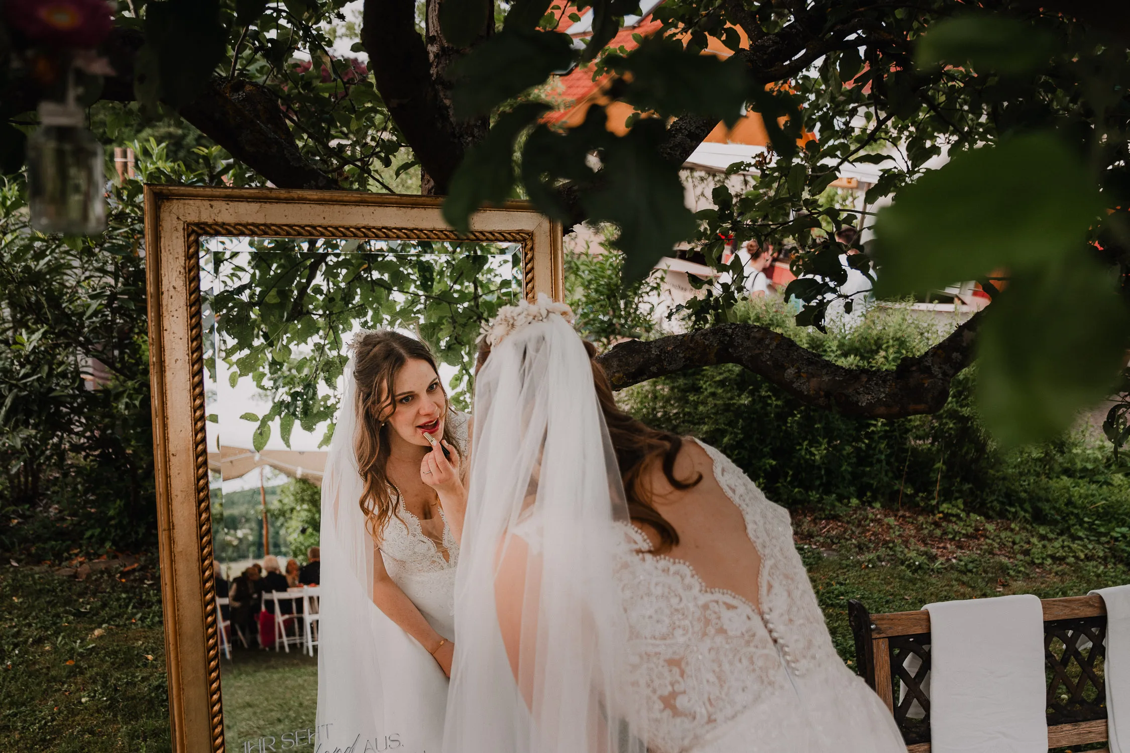 Hochzeitsfotograf Wuerzburg 23 Eine Braut in weißem Kleid und Schleier trägt Lippenstift auf, während sie in einen großen, verzierten Spiegel im Freien blickt, umgeben von Grün und Bäumen - die perfekte Inspiration für jede Gartenhochzeit, perfekt eingefangen von einem Hochzeitsfotografen Würzburg.