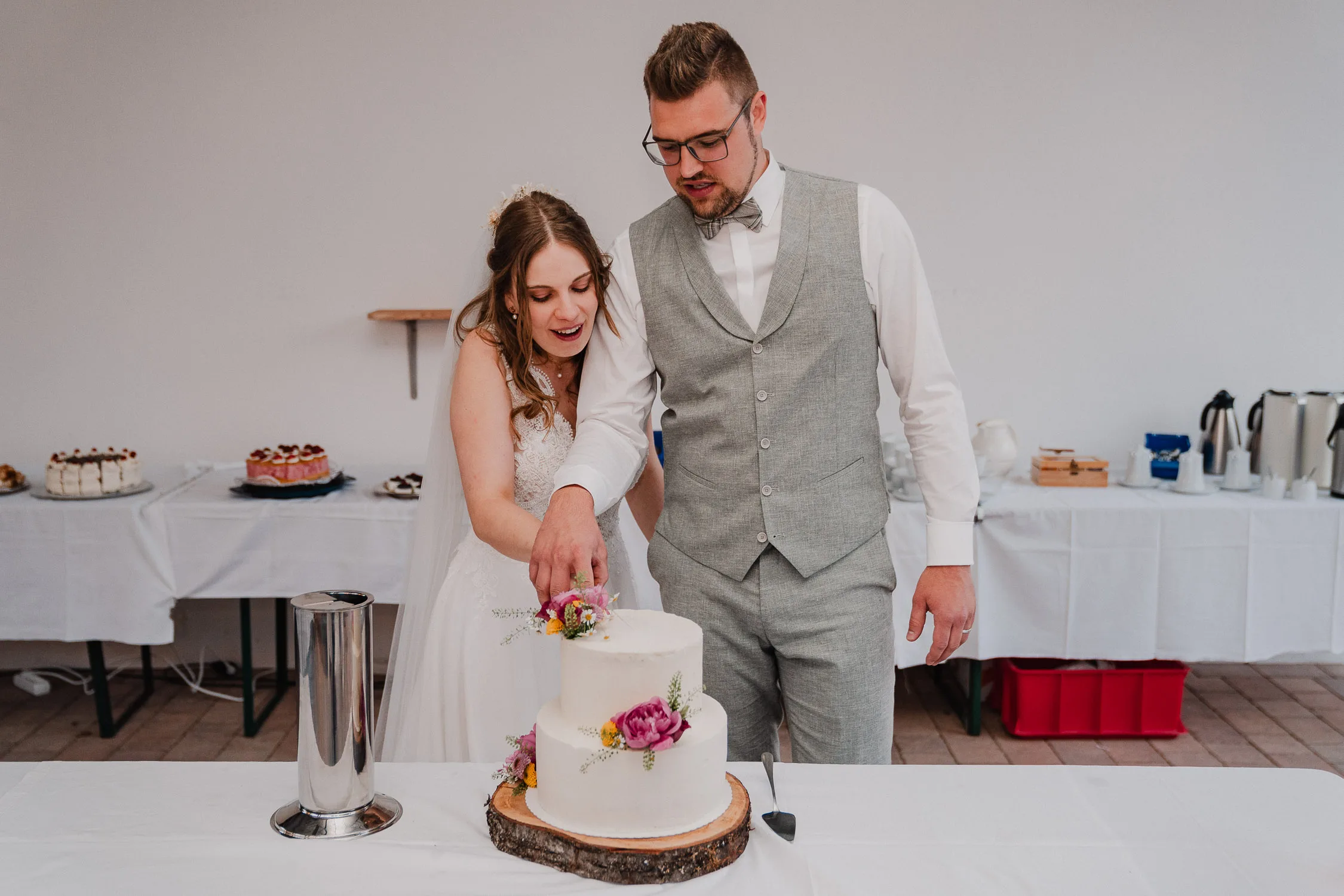 Hochzeitsfotograf Wuerzburg 24 Bei ihrer Gartenhochzeit schneiden die Braut und der Bräutigam eine weiße, zweistöckige und mit Blumen geschmückte Hochzeitstorte an. Das Paar steht hinter einem Tisch mit Desserts und Getränken und lächelt, während ein Fotograf diesen fröhlichen Hochzeitsmoment festhält.