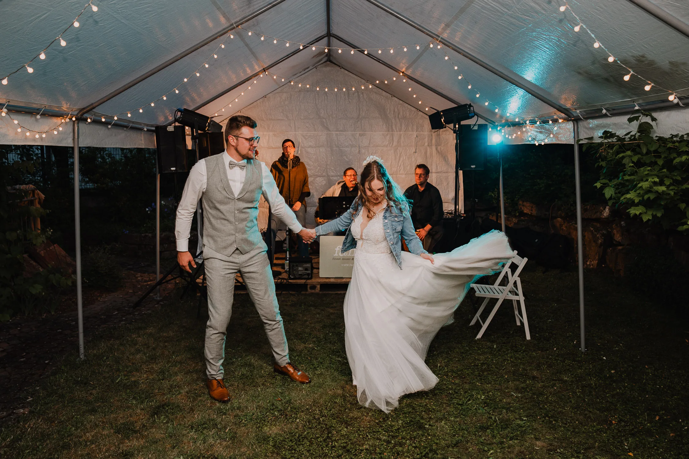 Braut und Bräutigam tanzen unter einem weißen, mit Lichterketten geschmückten Zelt auf ihrer Hochzeit. Der Hochzeitsfotograf Würzburg hält sie fest - den Bräutigam im grauen Anzug und Fliege, die Braut im weißen Kleid und Jeansjacke -, während im Hintergrund Musiker der Hochzeitsband "Famos" aus Karlstadt spielen.