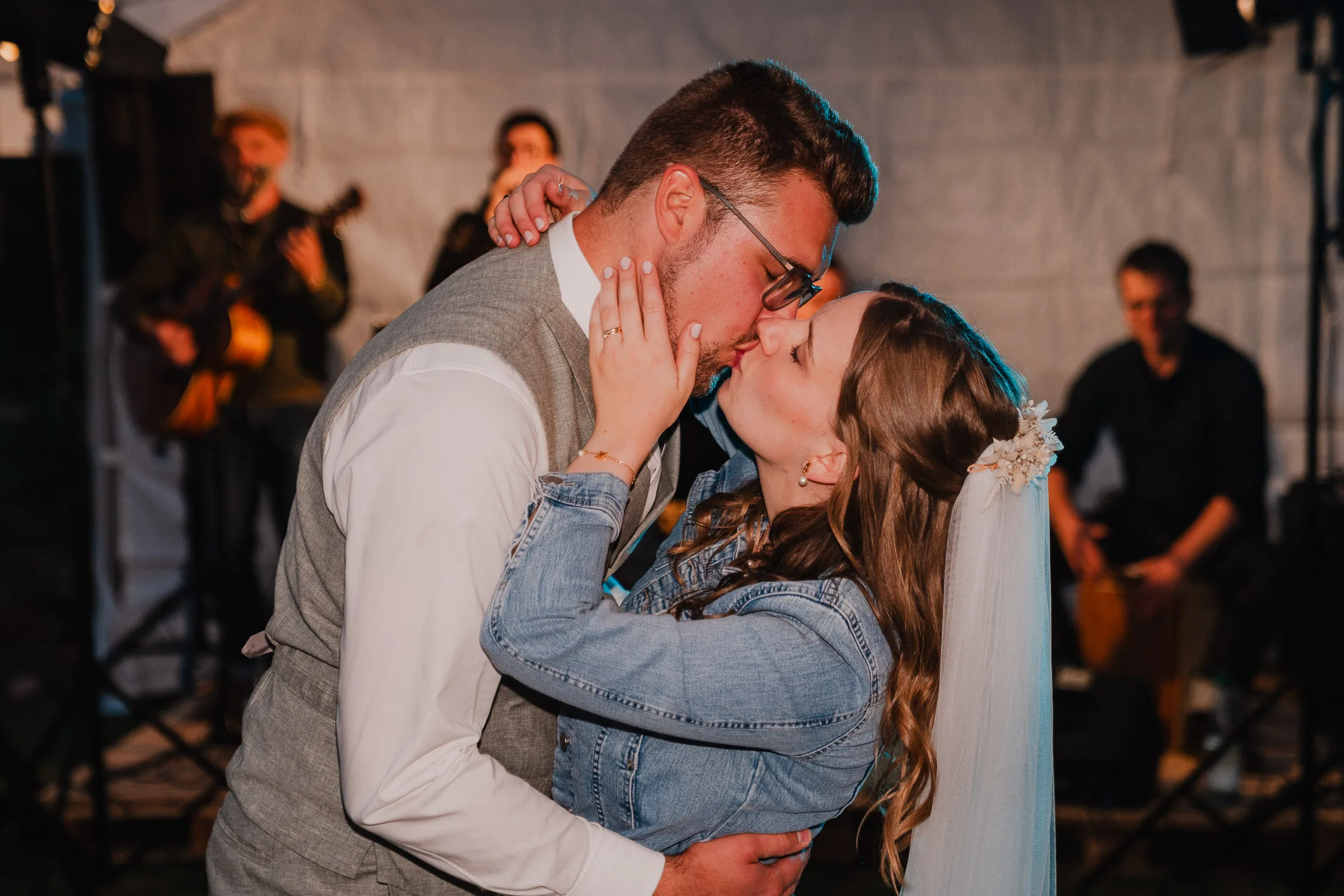Braut und Bräutigam geben sich während ihrer Gartenhochzeit einen romantischen Kuss auf der Tanzfläche, während im Hintergrund Musiker spielen. Die Braut trägt einen Schleier und eine Jeansjacke, der Bräutigam eine Weste und eine Brille - fotografiert von Hochzeitsfotograf Würzburg.
