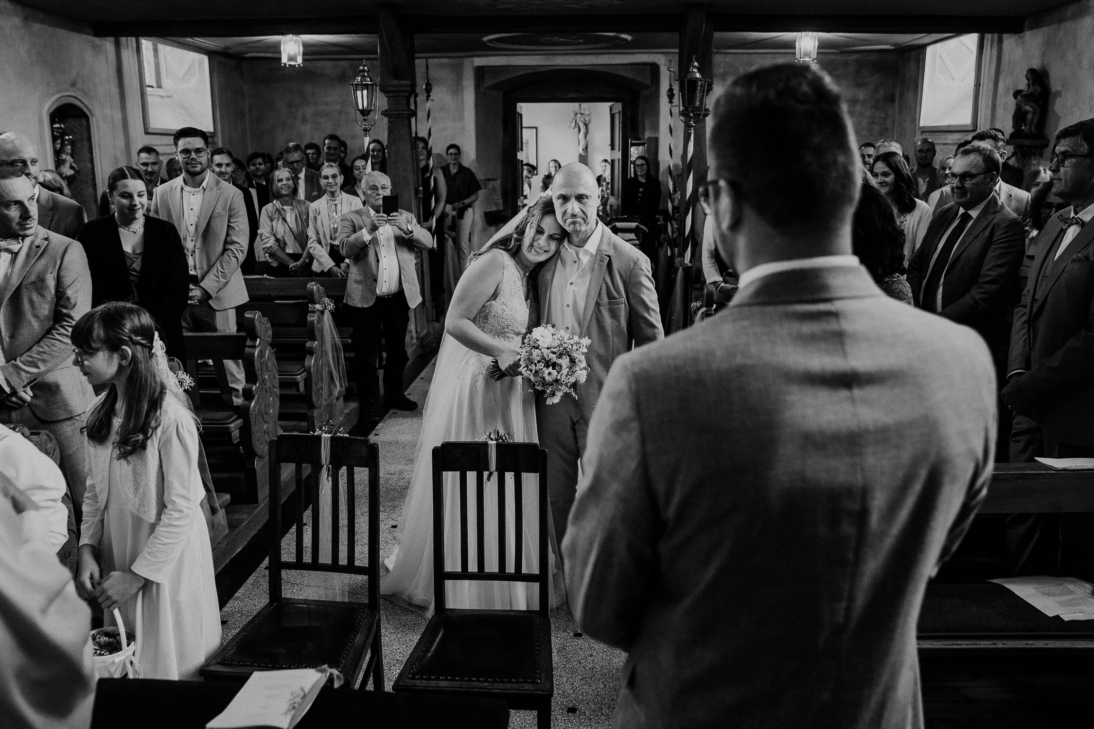 Eine Braut, die einen Blumenstrauß in der Hand hält, schreitet mit ihrem Vater lächelnd den Gang entlang. Die Gäste stehen und beobachten die Hochzeit in einer Kirche, während der Bräutigam im Anzug ihnen gegenübersteht - eine freudige, emotionale Szene für jeden Hochzeitsfotografen Würzburg.