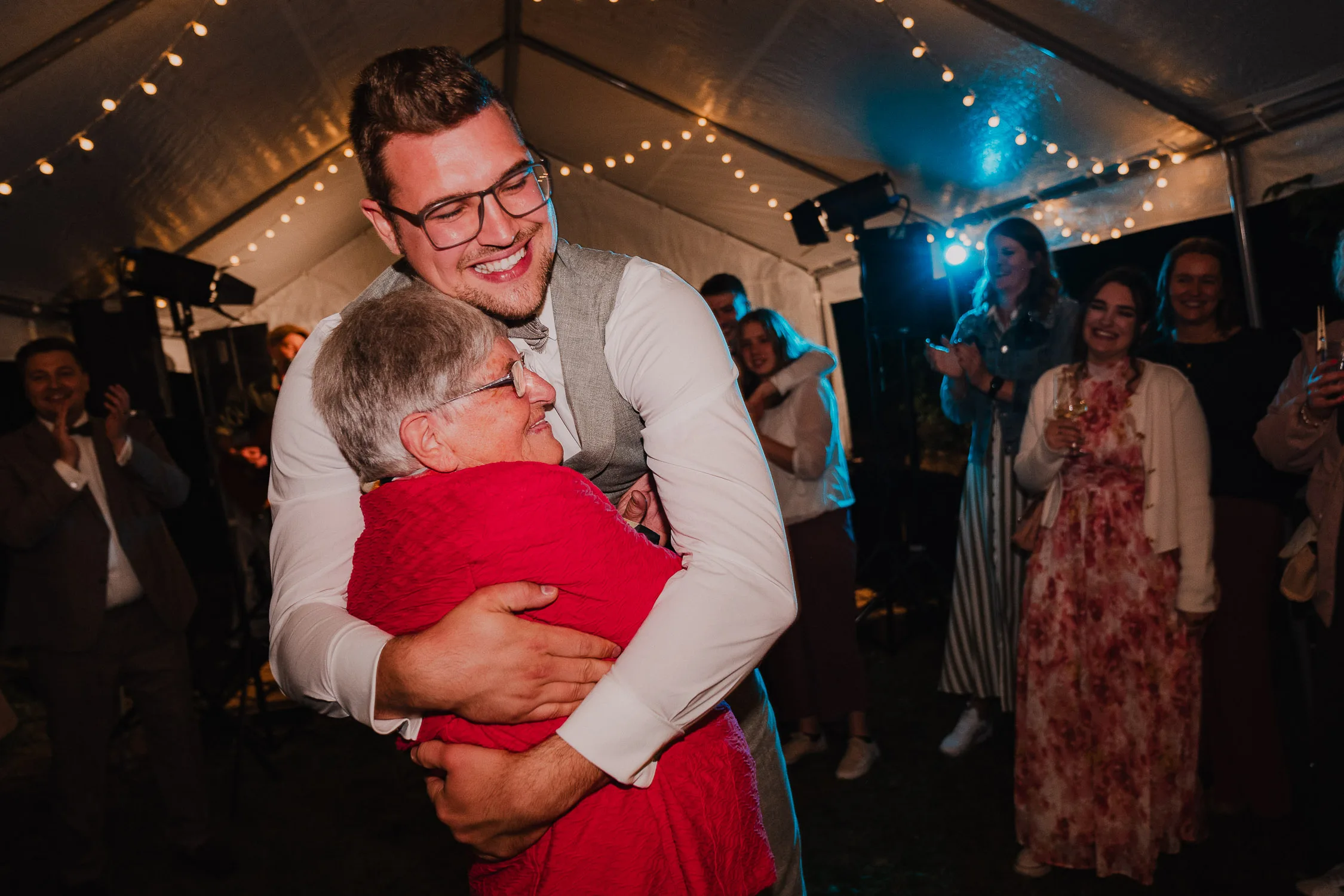 Ein junger Mann in einer Weste umarmt eine ältere Frau in einer roten Jacke unter einem Zelt mit Lichterketten, umgeben von lächelnden und klatschenden Gästen bei einer Hochzeit, aufgenommen von einem talentierten Hochzeitsfotografen Würzburg.