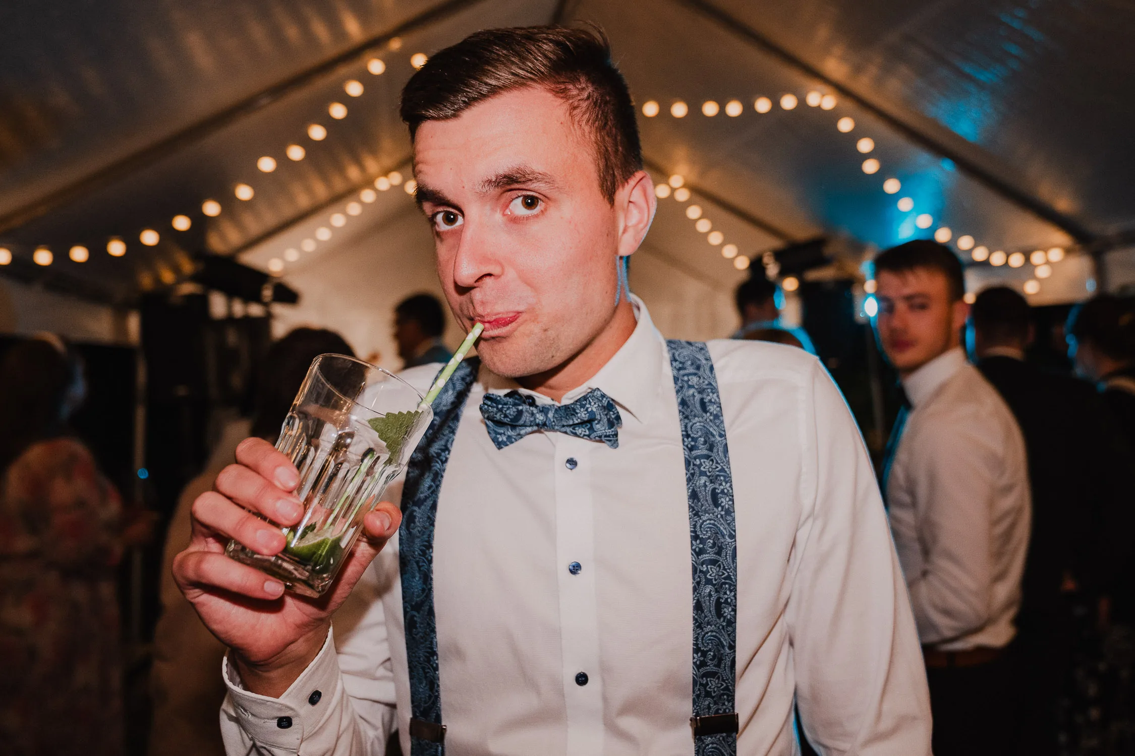Ein Mann in weißem Hemd, Fliege und Hosenträgern trinkt einen Cocktail mit einem Strohhalm bei einer festlich beleuchteten Hochzeit und schaut dabei in die Kamera. Aufgenommen von einem Hochzeitsfotograf Würzburg, schaffen Lichterketten und Gäste eine fröhliche Atmosphäre.