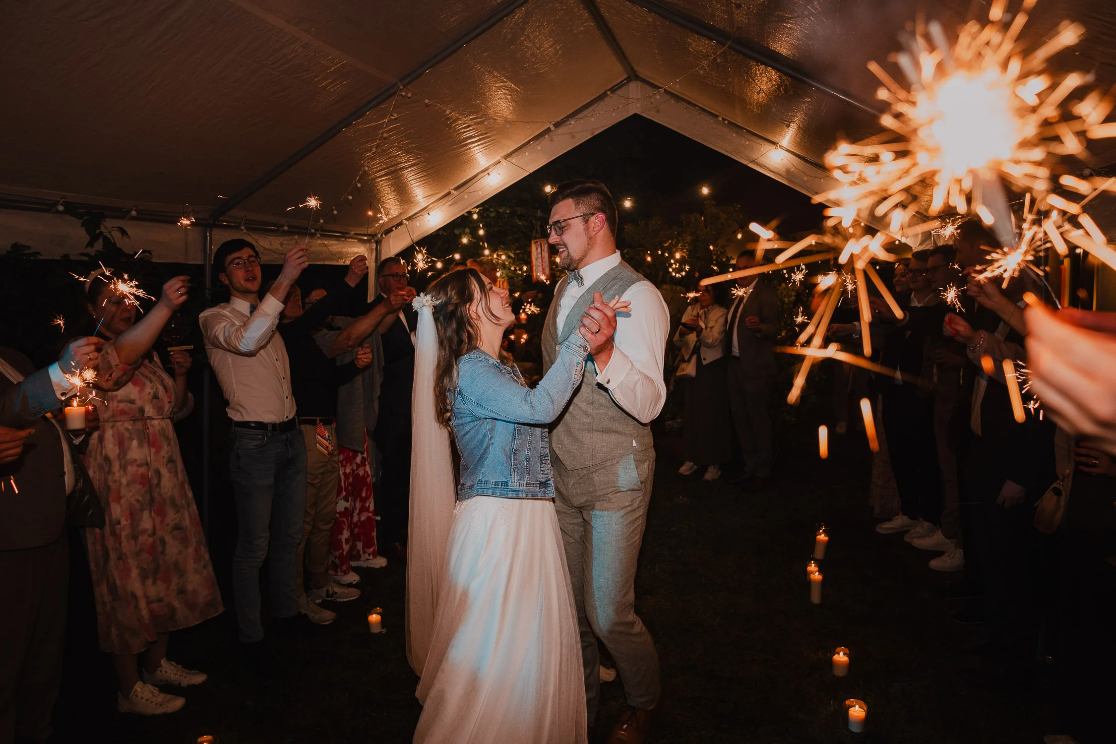 Braut und Bräutigam tanzen nachts unter einem Zelt, umgeben von Gästen, die Wunderkerzen und Kerzen auf den Boden stellen. Hochzeitsfotograf Würzburg hält diese fröhliche Hochzeit fest, bei der alle lächeln und in festlicher Atmosphäre feiern.