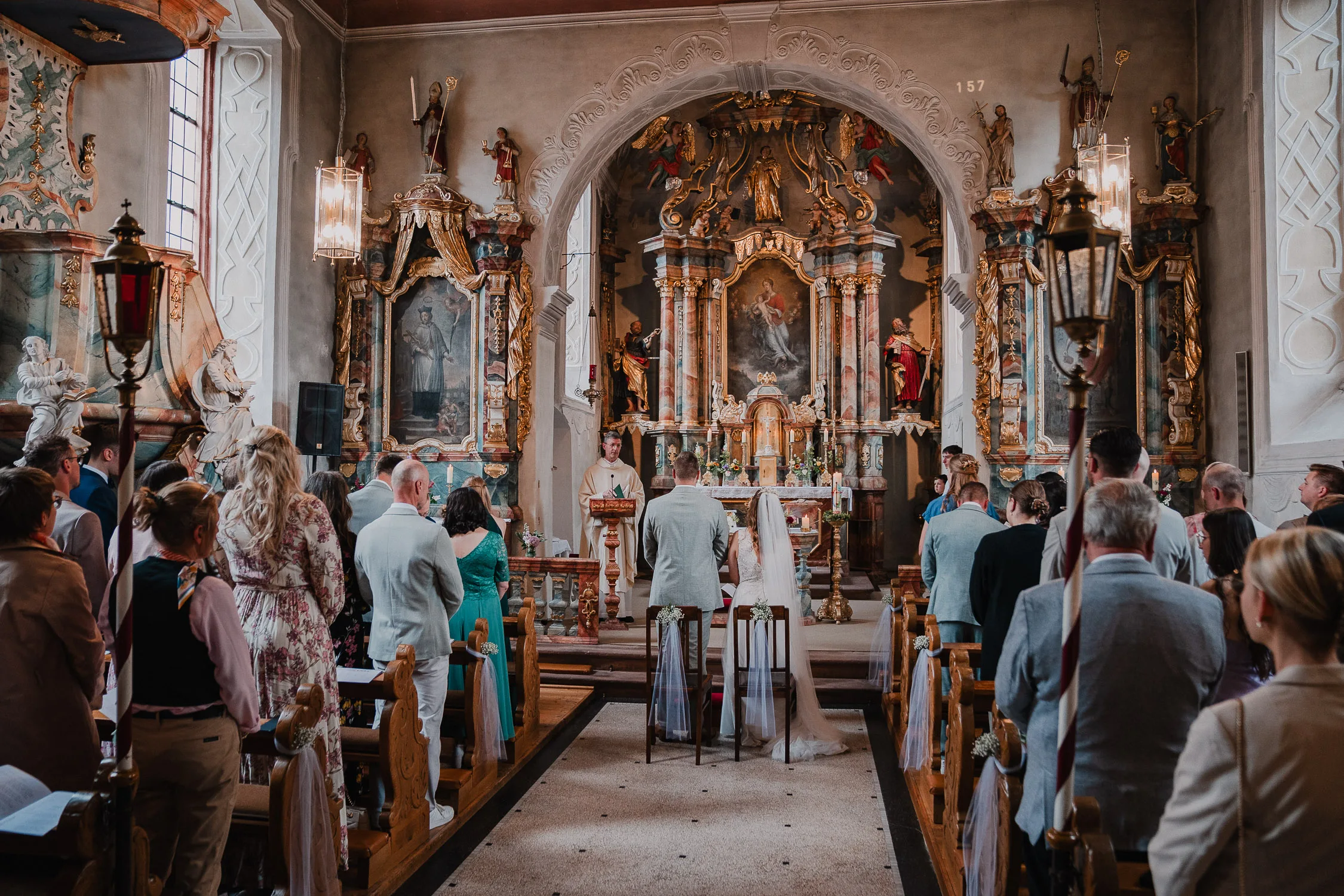 Braut und Bräutigam stehen während einer Hochzeit in einer kunstvoll geschmückten Kirche am Altar, umgeben von Gästen, die in den Kirchenbänken sitzen, mit kunstvollen Kunstwerken und Statuen im Hintergrund - perfekt für einen Hochzeitsfotografen Würzburg.