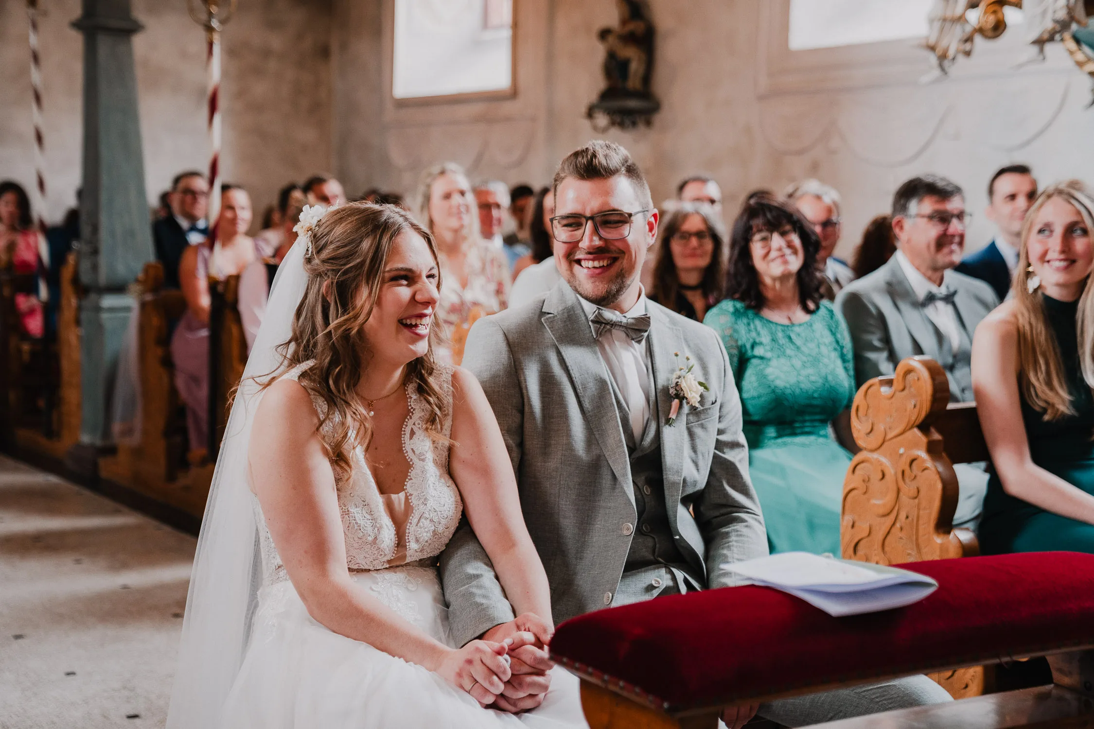 Braut und Bräutigam sitzen während ihrer Hochzeit lächelnd und händchenhaltend vor einer Kirche, umgeben von Gästen, die ebenfalls lächeln und ihnen zusehen - perfekte Momente für einen Hochzeitsfotografen Würzburg.