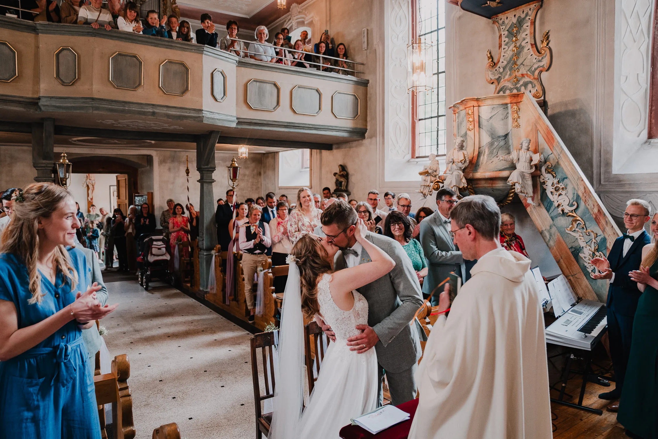 Ein frisch verheiratetes Paar küsst sich vor dem Altar in einer Kirche, umgeben von applaudierenden Gästen. Die Braut in Weiß und der Bräutigam in Grau sonnen sich im Sonnenlicht, das durch die Buntglasfenster fällt.
