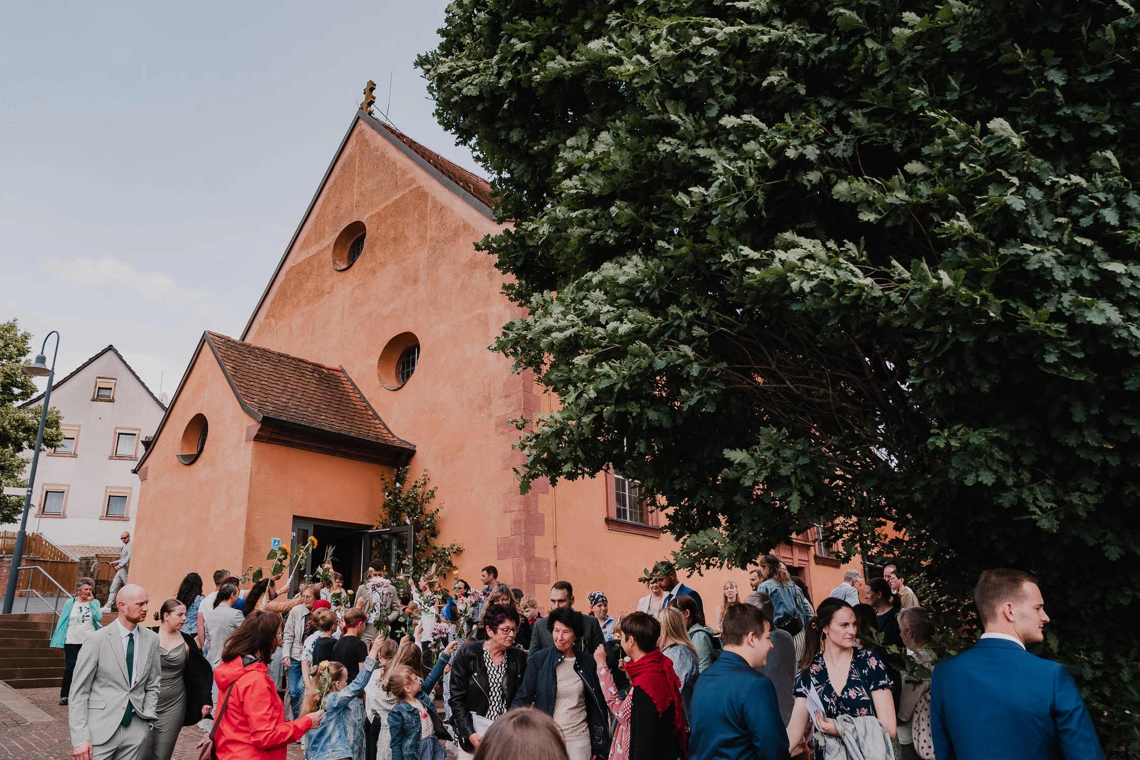 Eine große Gruppe von Menschen, gekleidet in formeller und halbformeller Kleidung, versammelt sich vor einem rustikalen orangenen Gebäude mit runden Fenstern, eine Kirche - aufgenommen von einem Hochzeitsfotografen Würzburg bei einer schönen Hochzeit. Grüne Bäume und Dorfhäuser vervollständigen die Szene.