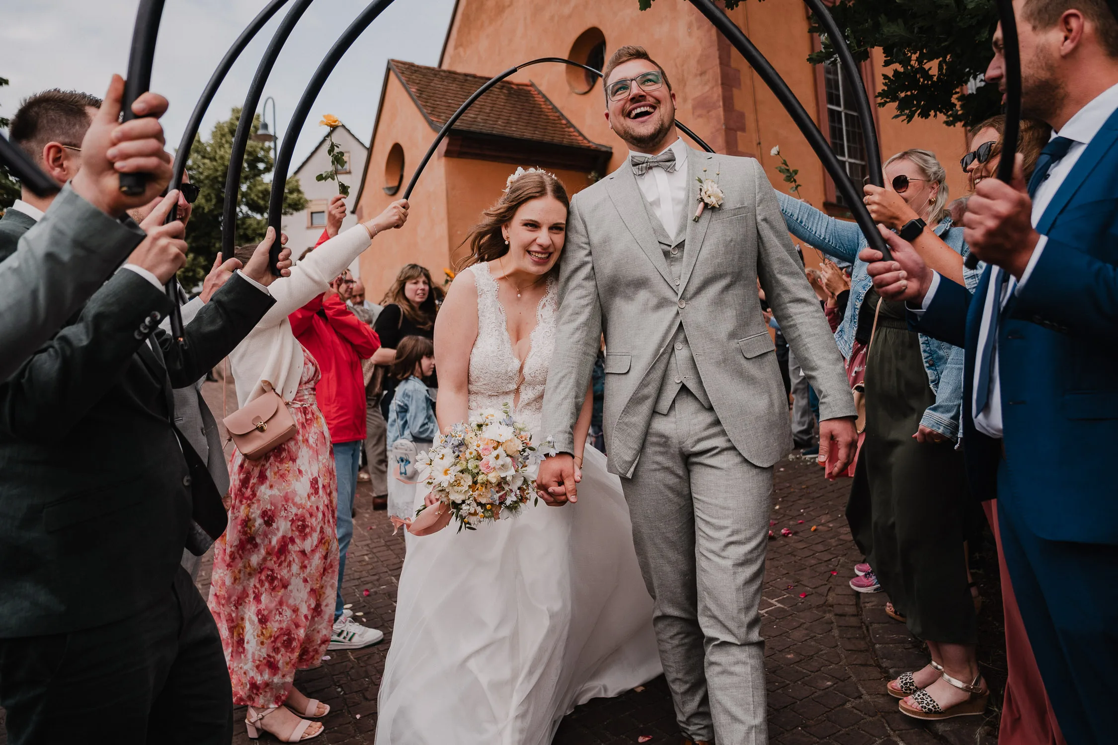 Ein lächelndes Brautpaar schreitet Hand in Hand unter einem Torbogen aus schwarzen Stangen, gehalten von Gästen vor einer Kirche, während die Menge jubelt und Fotos macht - wunderschön eingefangen von Hochzeitsfotograf Würzburg auf dieser Hochzeit.
