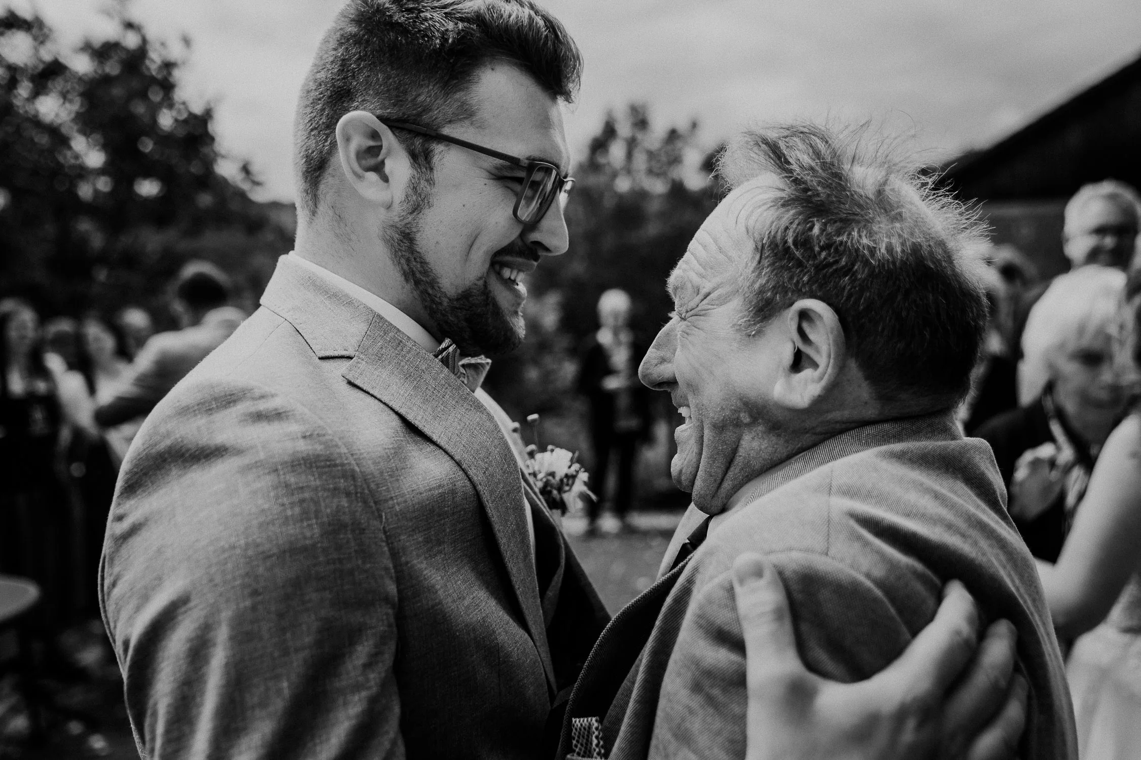 Ein junger Mann im Anzug lächelt warm, als er einen älteren Mann auf einer Hochzeit umarmt. Beide wirken fröhlich, umgeben von unscharfen Menschen bei einer Veranstaltung im Freien. Das Schwarz-Weiß-Foto fängt einen ehrlichen Hochzeitsfotograf Würzburg Moment ein.