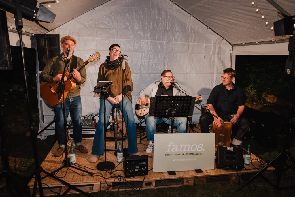 Vier Musiker der Hochzeitsband "Famos" treten auf einer kleinen Freiluftbühne unter einem weißen Zelt bei einer Gartenhochzeit auf. Zwei spielen Gitarren, einer singt und einer spielt Schlagzeug. Ein Schild mit der Aufschrift famos. steht davor, perfekt für jeden Fotografen oder Hochzeitsfotograf Würzburg, um den Moment festzuhalten.